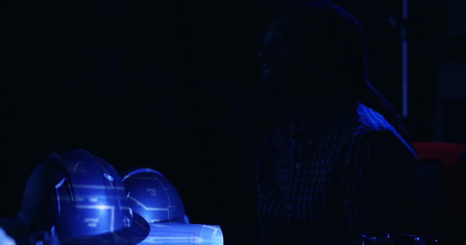 Two hard hats sit on table in a dark room with the reflection of a blueprint projector on them and a person in the shadows points and gestures behind them.