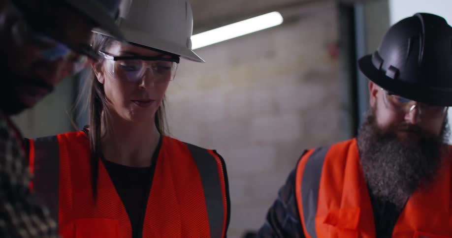 View panning left to right of three construction workers, a black male, a white female, and a bearded white male, standing in an indoor construction zone and collaborating on a project.
