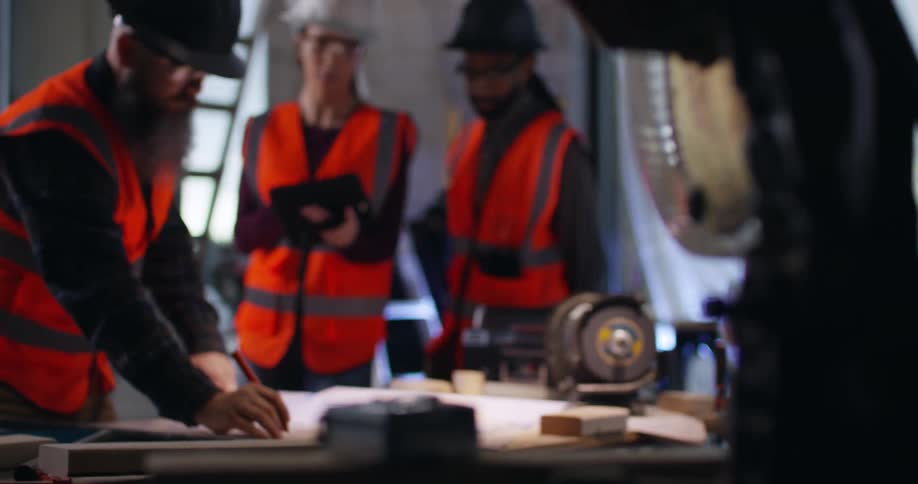 Three construction workers, a woman and two men, wearing orange reflective vests, eye protection, and hard hats collaborating over project plans in an active construction area.