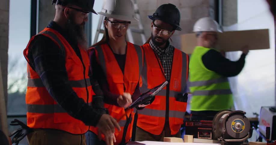 View panning forward through construction equipment of an active building site with a female and two male civil engineers working on a project and comparing notes in the foreground.