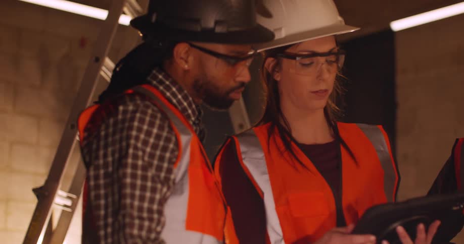 Three diverse workers wearing hard hats and safety gear are standing in a construction site at night and having a discussion on the project plans they are viewing on a tablet.