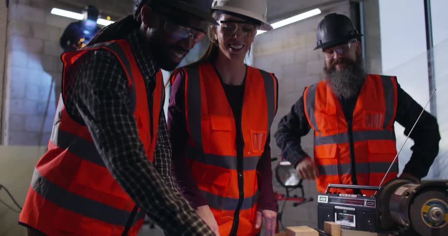 A female and two male construction workers in full safety gear gesture excitedly over blueprints laid out on a wood surface in an active indoor construction zone while a welder throws sparks while working on a task in the background.