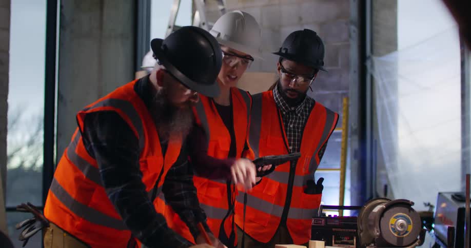 A bearded white male foreman in an active construction site bends over taking notes on project plans as he is approached by two others, a woman and a man, who gesture to a tablet and compare notes.