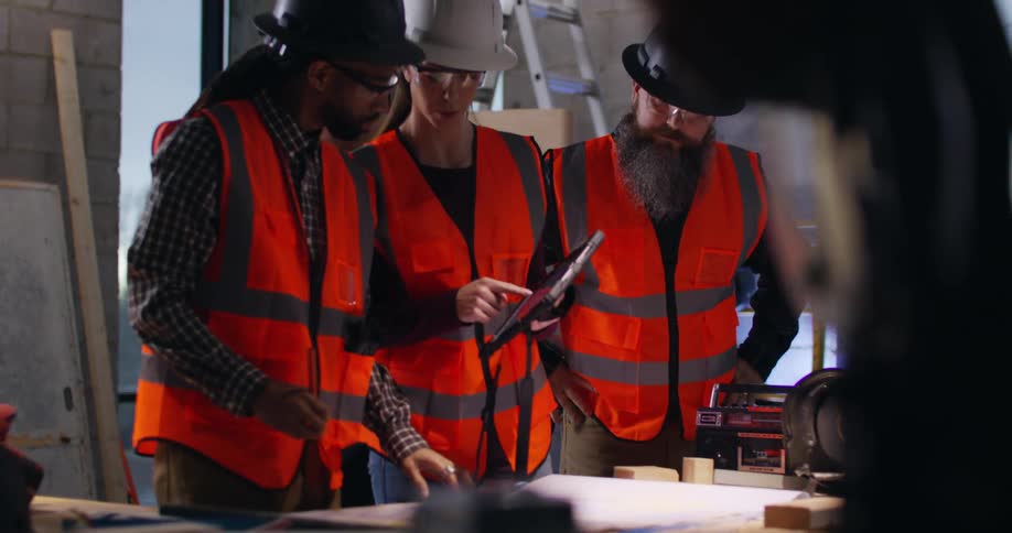 POV from behind a saw of 3 construction workers, a woman and two men wearing safety gear and hard hats, discussing and making notes over project plans in an active indoor building site.