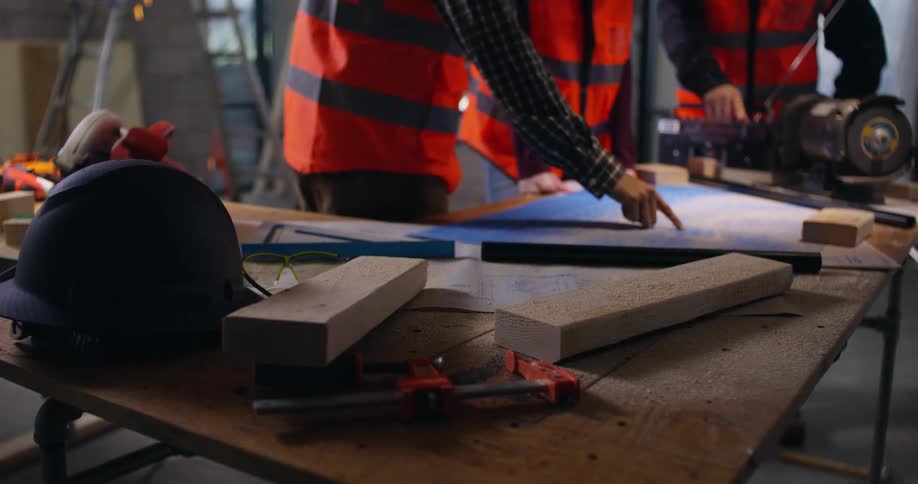 Hands of three construction workers in orange safety vests working with blueprints on a table covered in tools, two-by-four pieces of wood, and sawdust at an active building site with welding sparks falling in the background.