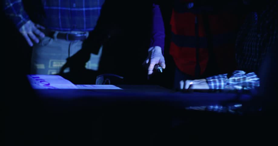Two people standing in a dark room lit only by the blue light of a screen pointing out details on a paper spread on a table.