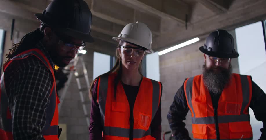 A female and two male construction workers, wearing safety gear and hard hats, are standing in an active indoor construction site discussing project plans while a welder works on a ladder behind them.