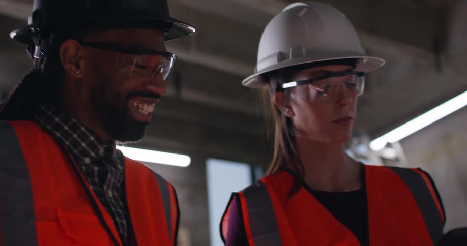 A female civil engineer makes notes on a tablet while talking to a black male foreman in a building under construction.