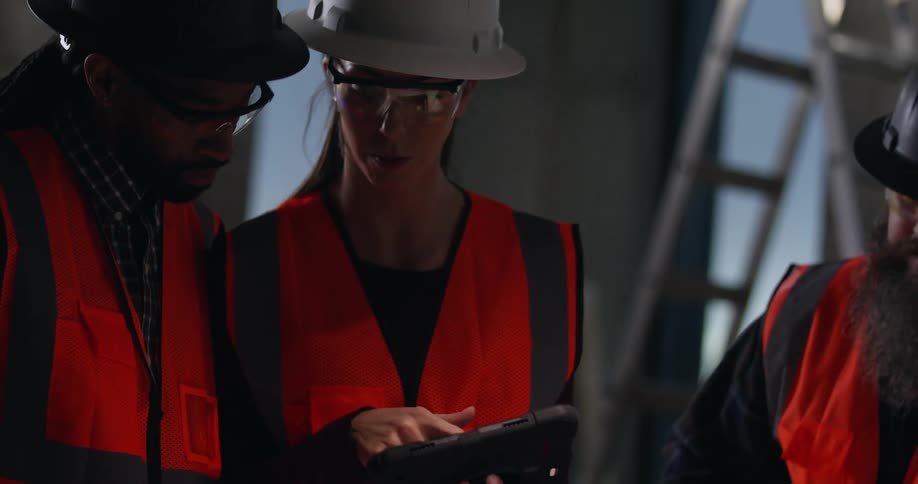 A woman and two men, wearing safety gear in an active construction site, discuss and analyze project blueprints while making notes on a digital device.