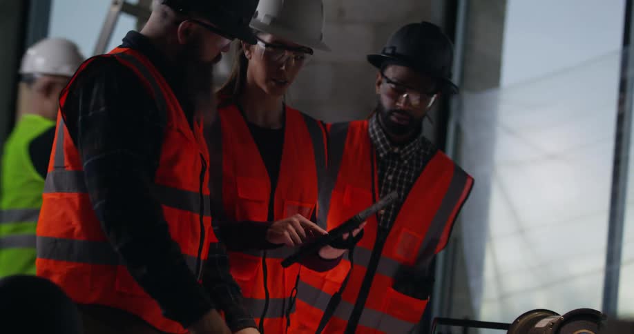 A bearded, white male foreman stands in an active construction site taking notes and examining blueprints, as two supervisors, a white woman and a black man, approach him and have a discussion over the building plans as welding sparks fly behind them.