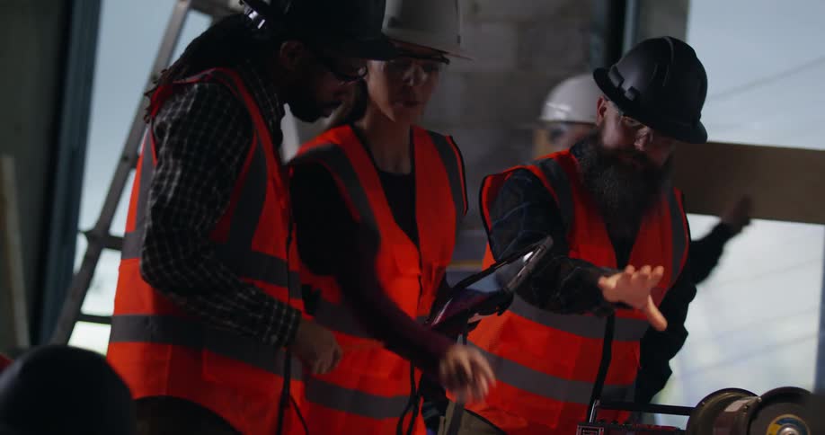 A group of diverse construction workers wearing safety gear discuss project plans with the foreman in an active indoor building site as other workers cross behind and welding sparks fly.