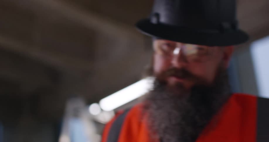 Close up of construction workers, a female and bearded male, standing in an active building construction area wearing safety gear and hard hats and interacting over blueprints.