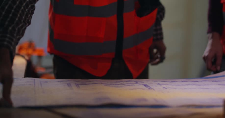 Two construction workers are at a table full of saw dust on a job site when one picks up a set of blueprints and moves them away to show a different set below.