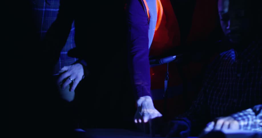 A female civil engineer wearing an orange safety vest and hard hat is pointing out details on blueprints to two men in a dark room illuminated by a projector.