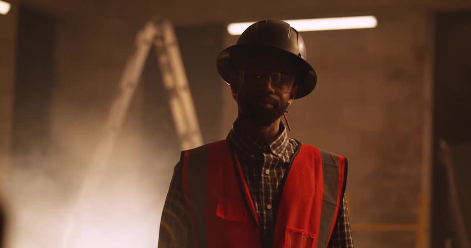 Portrait of a bearded, black male construction worker wearing a safety vest, eye protection, and a hard hat in a dusty and dimly lit indoor building site.