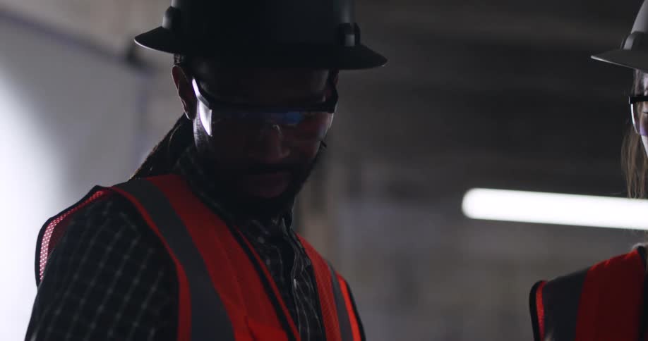 Two construction workers, a black male and white female, wearing safety vests, eye protection, and hard hats stand together in an indoor construction site and discuss blueprints and building plans.