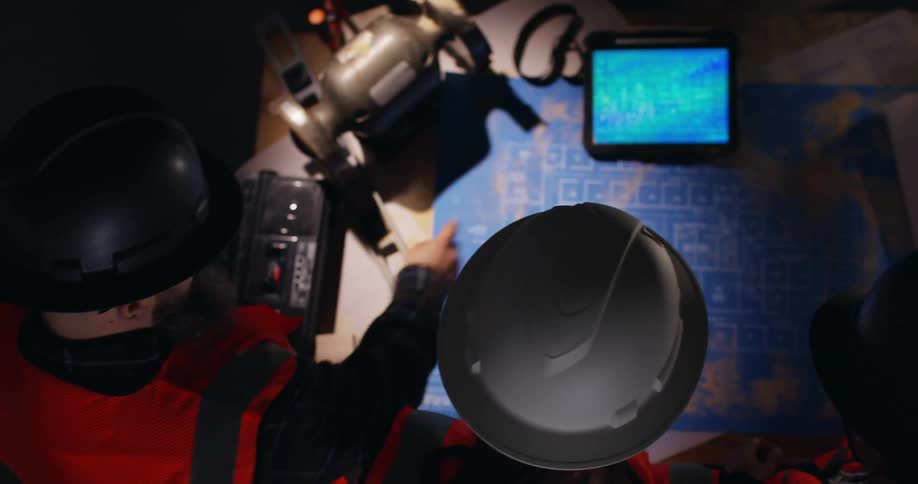 View looking down on three engineers in protective gear at a construction site analyzing both printed project blueprints and digital ones on a screen.