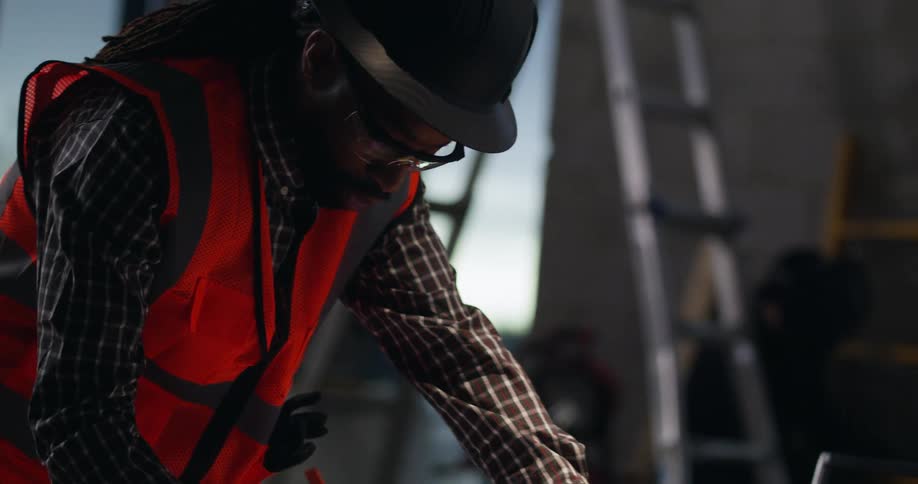 A black male foreman in hard hat, reflective vest, and safety glasses is standing in an active indoor high-rise building site looking down at and making notes on construction plans.