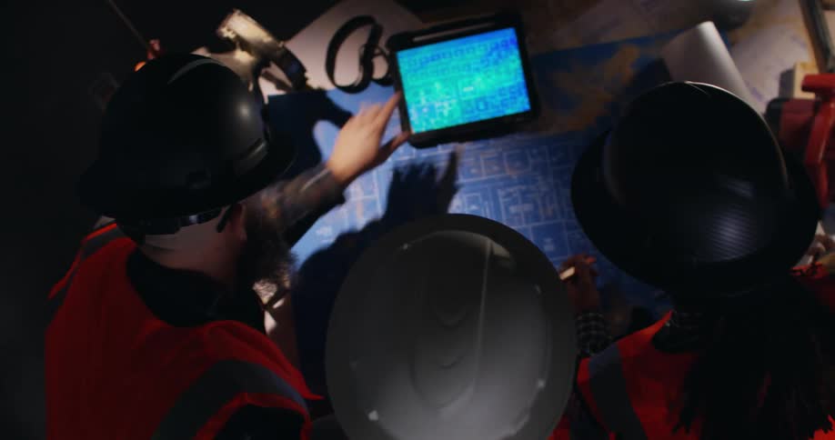 View looking down on three construction workers in protective hard hats and orange vests who are looking at both printed and digital blueprints on a tablet screen.