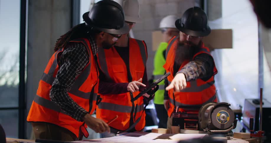 A black male civil engineer in an active construction area bends over taking notes on project plans as two others, a man and woman, join him to discuss details.