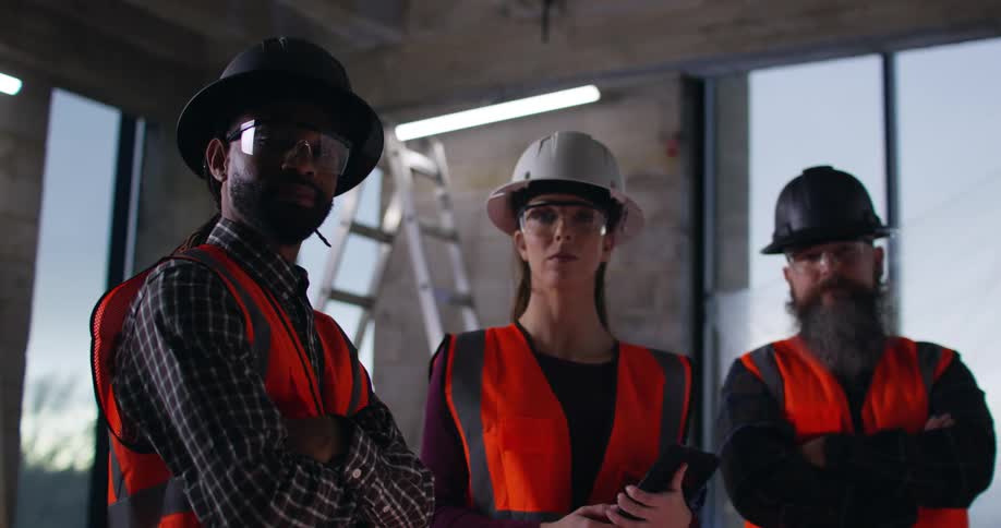 Portrait of a female and two male construction workers indoors at an active high-rise building site wearing safety vests, eye protection, and hard hats.
