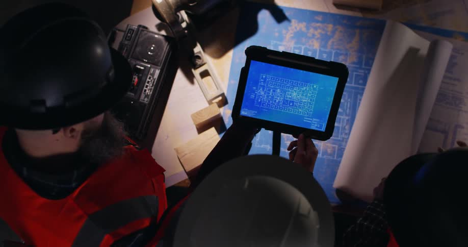 View from above of construction workers wearing hard hats and orange safety vests who are examining a digital tablet displaying a detailed blueprint with a large printed blueprint spread out on a table below.