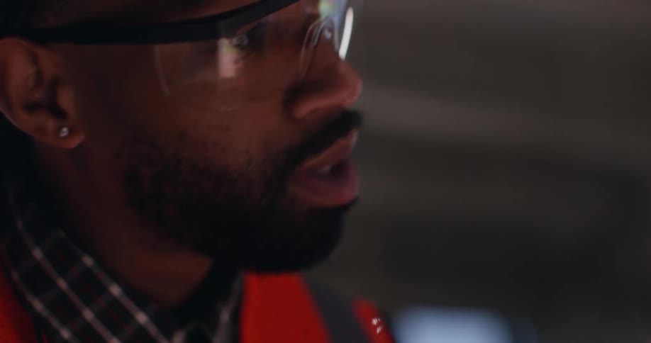Close up of two civil engineers, a bearded man of color and a white woman, both wearing safety goggles, reflective vest, and a hard hat, talking to each other in an active construction area.