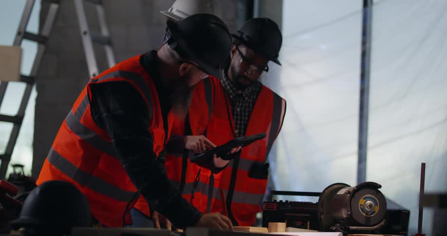 A construction project manager reviews building plans on a workbench at an active construction site as a pair of diverse engineers, a black male and white female, walk over to discuss.