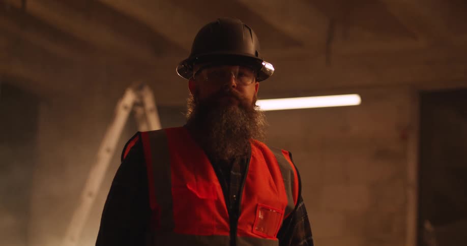 Portrait of a burly, bearded Caucasian construction worker standing with face shadowed an indoor building site wearing a hard hat, eye protection, and safety vest.