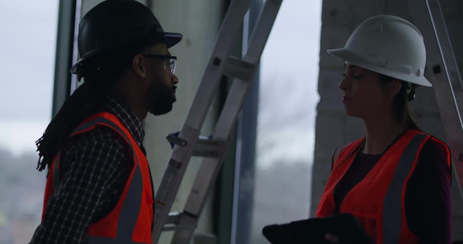 A black man and white woman wearing hard hats, safety vests and eye protection inside a construction site stand near a ladder and have a conversation.