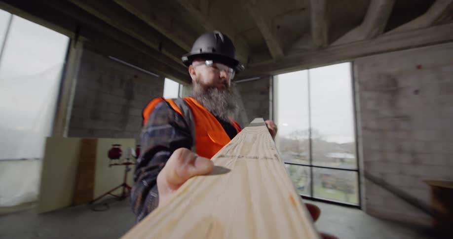 A bearded worker wearing an orange safety vest and hard hat in a building under construction makes a measurement on a piece of wood and then carries it over to a miter saw to make the cut.