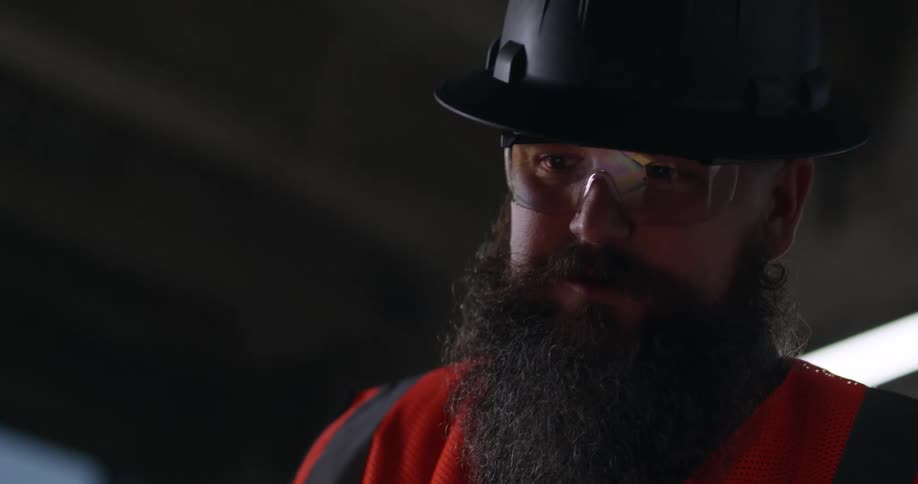 A burly, bearded man wearing a hard hat, safety vest, and eye protection at a construction site stands a discusses plans with someone off camera.