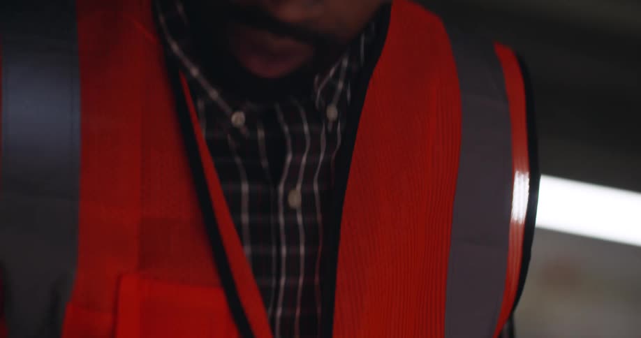 A black man with facial hair and dreadlocks, wearing a safety vest, eye protection, and a hard hat looks down at some construction plans and rubs his hands together as he discusses them with a partner.