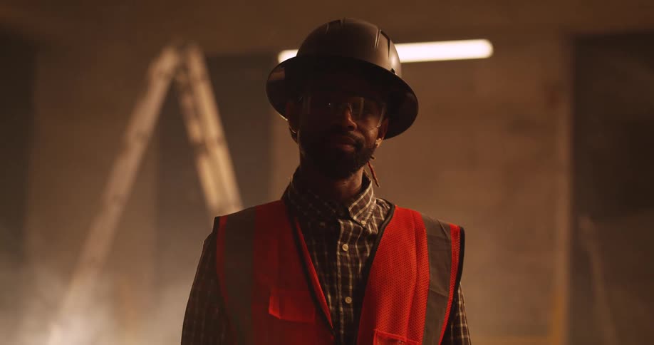 Portrait of a black, bearded male construction worker with protective eyewear, safety vest, and hard hat on at a dimly list and dusty building site.