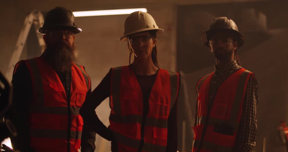 Portrait of three contractors; a woman, a black man, and a bearded white man, standing together in a dusty and dimly lit construction area with hard hats and safety gear on.