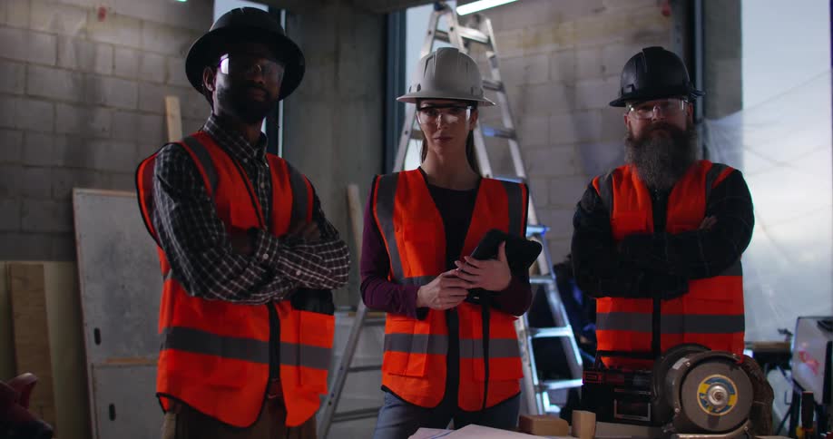 Three diverse contractors wearing safety gear stand in a construction zone in a high-rise with a ladder and sparks flying behind them.
