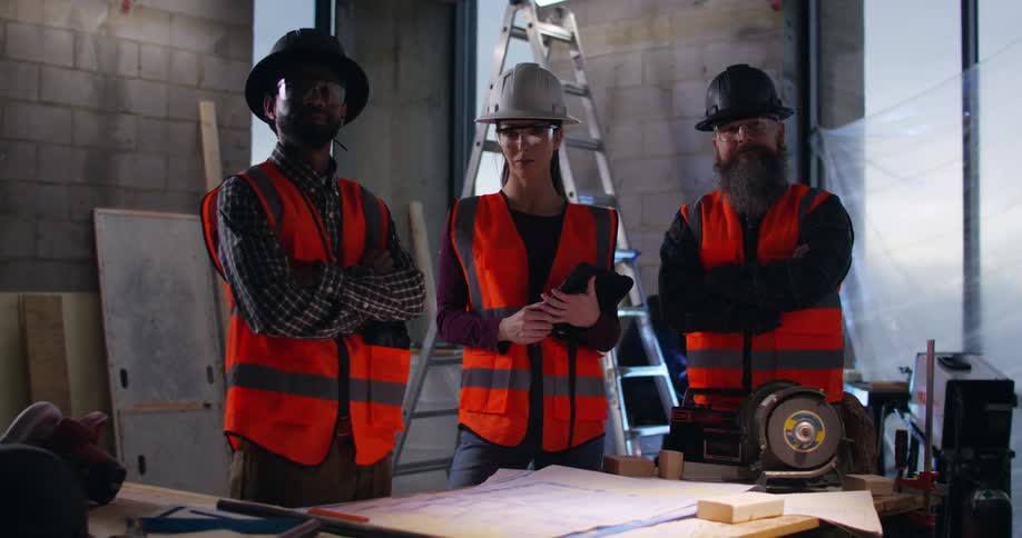 A diverse group of three construction workers wearing safety vests, eye protection, and hard hats stand indoors at a construction site with blueprints on the surface in front of them as sparks fly from a welding job behind them.