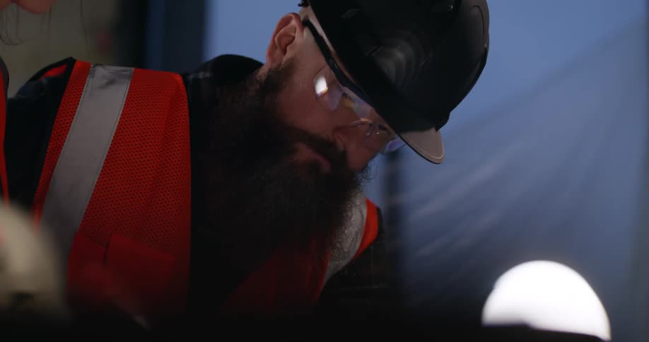 A bearded project manager wearing a safety vest, protective eye gear, and hard hat stands at an indoor construction site and looks down intently at building plans illuminated by a nearby light bulb.