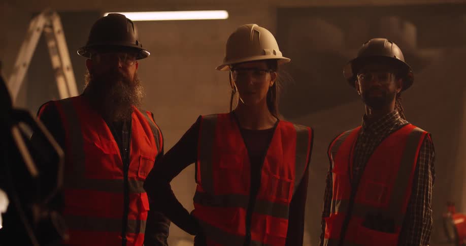 Portrait of a female and two male construction workers wearing safety vests, eye protection, and hard hats stand in the shadows of a dusty indoor building site.