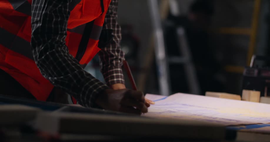 A bearded, black civil engineer with dreadlocks is making notes on project blueprints in an active indoor construction area.