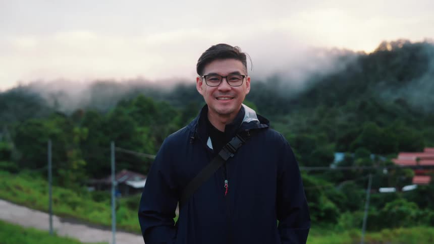 Standing on a hillside a smiling man gazes out over a rural village with a backdrop of misty mountains and a cloudy sky creating a peaceful and joyful scene as he takes in the expansive panoramic view from his elevated position