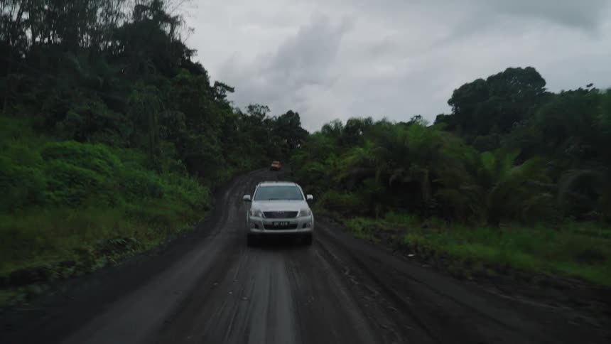 A silver suv drives along a dark dirt road winding through a lush green landscape under a cloudy sky