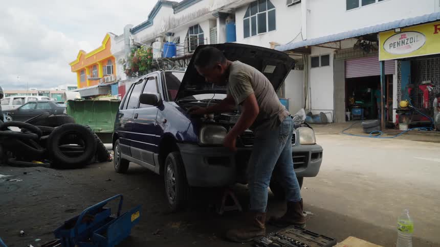 A mechanic is working on a blue car with its hood open in front of a row of shops with various tools and tires scattered around the area