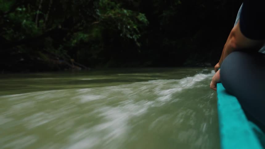 A firstperson perspective of the side of a teal boat speeding through a murky green river creating a white wake with dark foliage visible in the background