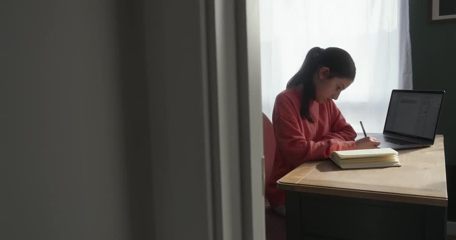 Girl sitting writing with a pencil next to an open laptop