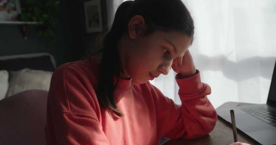A teen girl sitting at the table in front of a laptop while writing in a notebook