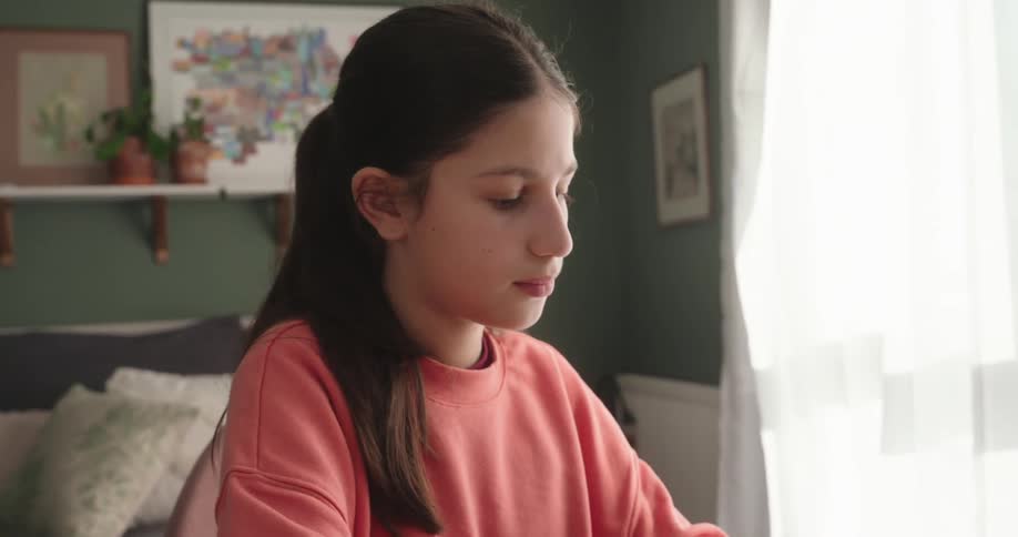 A girl wearing a pink sweatshirt is typing on a laptop in a room with a green wall and a window with white curtains