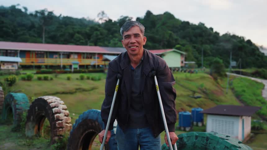 A person using crutches is standing outdoors on a path made of large painted tires with a background of buildings and lush hilly terrain