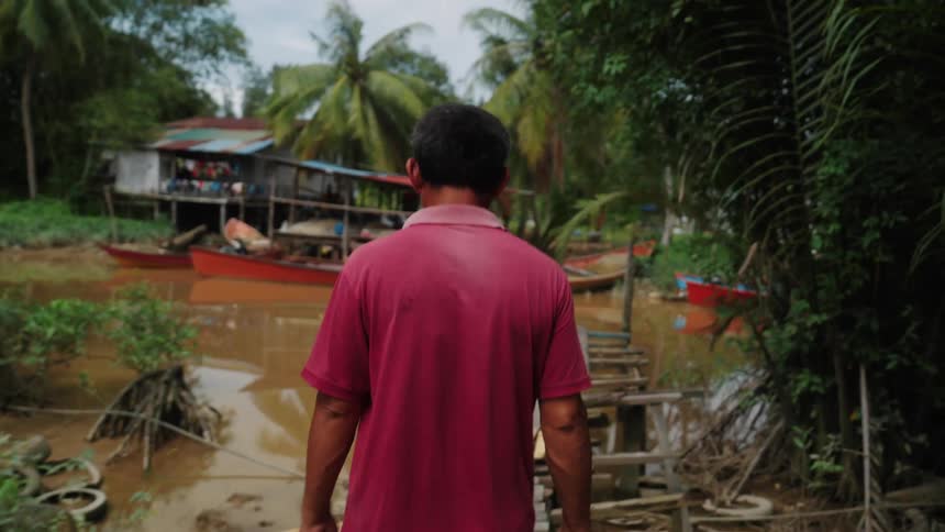 A person in a red shirt walks on a narrow wooden bridge over a muddy river with palm trees and a stilt house in the background