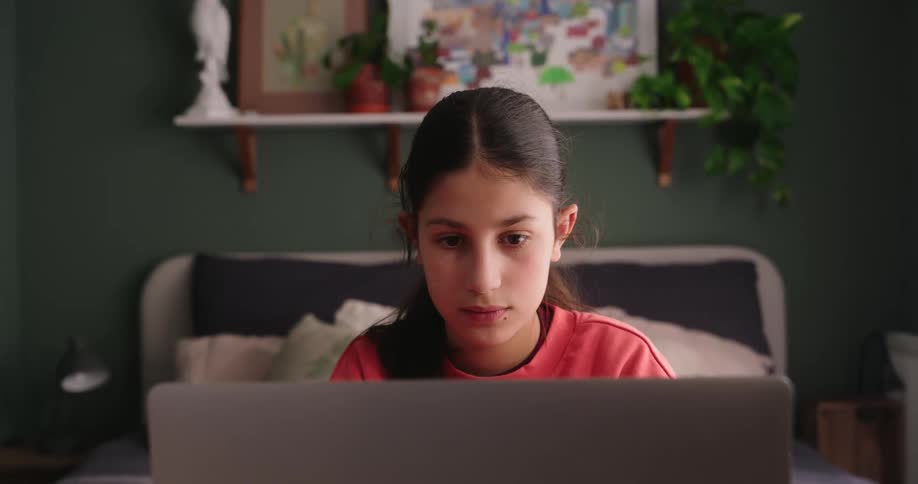 A teenage girl with long hair is studying on a laptop while at a desk in her bedroom